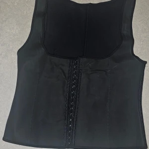 Svart träningsväst med snörning - Svart väst i syntetmaterial, perfekt för träning. Modellen är tight och har breda axelband samt hakar och snörning framtill för justerbar passform. Stilren och enkel design utan mönster, med slät yta och markerade sömmar.