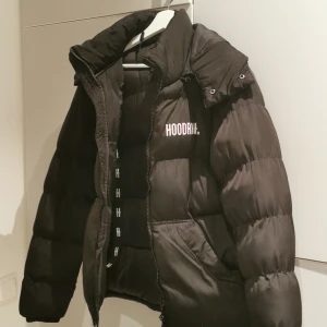 Svart pufferjacka från Hoodrich XS - Säljer en svart pufferjacka från Hoodrich i storlek XS. Jackan har hög krage, dragkedja framtill och logga på bröstet. Den är vadderad och har en klassisk pufferlook som är både snygg och varm. Perfekt för kalla dagar och streetstyle. Jackan är i xs men är större entligen. Köpt för ett och ett halv år sen. Luvan är också en dragkedja. 