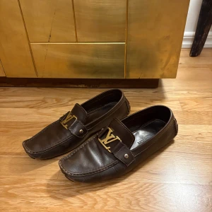 Bruna Louis Vuitton loafers i skinn - Snygga bruna loafers från Louis Vuitton i äkta skinn med guldigt LV-spänne framtill. Klassisk design med rund tå och detaljerade sömmar. Perfekta för dig som vill ha en stilren och lyxig look.