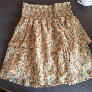 Blommig volangkjol från MSCH - Supersöt kort volangkjol från MSCH med blommigt mönster i orange, grön och gul på vit botten. Kjolen har smock i midjan och flera lager volanger som ger en luftig och somrig känsla. Perfekt för dig som gillar färg och mönster!