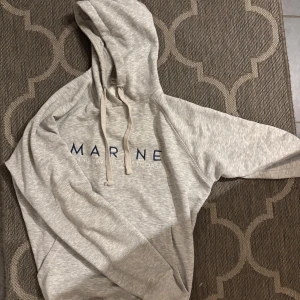 Hoodie Marine Scandinavian Westcoast S - Säljer en ljusgrå hoodie från Marine Scandinavian Westcoast med blå broderad logga på bröstet. Tröjan har huva med snörning och en klassisk känguruficka framtill. Perfekt för chill dagar och enkel att matcha med jeans eller mjukisbyxor. Storlek S