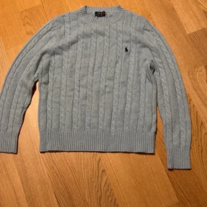 Ljusblå kabelstickad tröja Polo Ralph Lauren - Kabelstickad tröja från Polo Ralph Lauren i ljusblått med klassisk rund hals och broderad logga på bröstet. Tröjan är stickad i mjuk ull och har ribbade muddar vid ärmslut och nederkant. Perfekt för lager-på-lager under kyliga dagar.