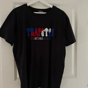 Svart Trapstar t-shirt med broderi - Svart t-shirt från Trapstar med broderad text i blått, rött och vitt på bröstet. Klassisk rund hals och korta ärmar. Materialet är mjuk bomull och passformen är normal. Perfekt för dig som gillar streetwear och vill sticka ut med snygga detaljer.