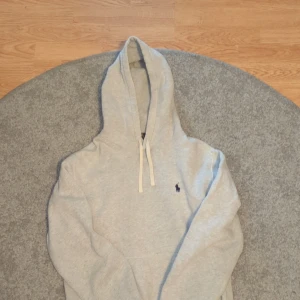 Grå hoodie från Polo Ralph Lauren - Ljusgrå hoodie från Polo Ralph Lauren med klassisk broderad logga i mörkblått på bröstet. Tröjan har huva med snörning och är tillverkad i mjuk bomullsmix. Perfekt för chill dagar och snygg till jeans eller joggers. Den är använd väl men inga stora defekter. Var inte rädda att fråga om fler bilder. Priser är inte hugget i sten!