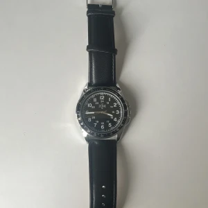 Svart armbandsur från InZone 47 - Stilren klocka från InZone 47 med svart urtavla och tydliga vita siffror. Boetten är i silverfärgad metall och armbandet är svart med ljus insida, Klockan är helt ny och inte använd en ända gång. Eftersom fick den i present men säljer den eftersom de inte är min stil.