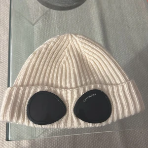 Vit ribbstickad mössa C.P. Company - Vit ribbstickad mössa från C.P. Company med klassiska svarta goggles framtill. Mössan är i grovstickad bomull och har en uppvikt kant. Perfekt accessoar för att sticka ut och hålla värmen under kyliga dagar. Ny haft den i en vecka kvitto finns 