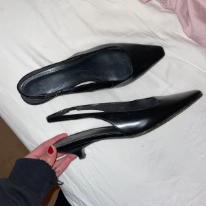 Svarta klackar  - Snygga svarta pumps från H&M med spetsig tå och låg klack. Skorna har en öppen häl med smal rem och är tillverkade i blankt skinnliknande material. Använda 1 gång 