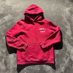Röd hoodie från In Gold We Trust - Säljer en snygg röd hoodie från In Gold We Trust med vit logga tryckt på bröstet och ryggen. Tröjan har huva med tryck, känguruficka och ribbade muddar. Perfekt för dig som vill ha en streetstyle-look med attityd.