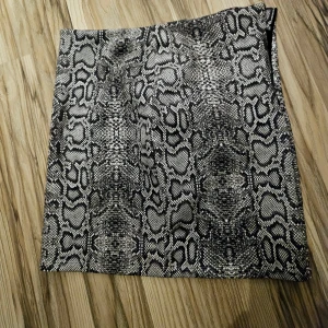 Kort kjol med ormmönster - Snygg kort kjol med svart och grått ormmönster. Kjolen har en rak och tight passform som ger en cool vibe. Dragkedja på sidan . Materialet känns som syntet och är lätt att bära. Perfekt för dig som gillar att sticka ut med djurmönster.