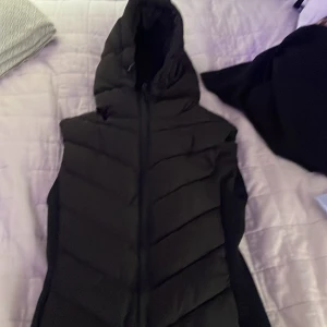 Svart dunväst med huva från Zara - Svart dunväst från Zara i storlek XS med quiltad design och dragkedja framtill. Västen har en stor huva och är perfekt för lager-på-lager-stil. Snygg och enkel att matcha med olika outfits.
