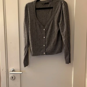 Grå kofta från ONLY med knappar - Grå kofta från ONLY med v-ringning och långa ärmar. Koftan har en figurnära passform och stängs med vita knappar framtill. Perfekt att slänga över en topp eller klänning för en chill look.