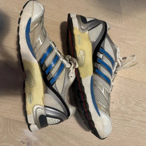 Adidas Supernova sneakers blå/vit - Adidas Supernova sneakers i mesh och syntet med klassiska tre blå ränder på sidorna. Skorna är vita med inslag av grått, blått och svarta detaljer samt färgglad sula med rött och blått. Platt sula och snörning för bra passform. Perfekta för träning eller löpning.
