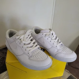 Nike SB vit sneaker low - Fräscha vita Nike SB sneakers med lågt skaft och klassisk design. Skorna har perforerad tå, snörning och är tillverkade i skinn med mjuk vadderad plös och häl. Snyggt Swoosh-märke på sidan och stilren look som passar till streetwear.