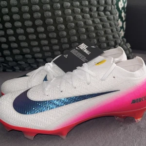 Nike Mercurial Air Zoom fotbollsskor - Nike Mercurial Air Zoom fotbollsskor i vit med rosa och röd gradient på sidorna. Swoosh-logga i metallicblå, snörning och låg profil. Sulan har röda och vita detaljer med dobbar för gräsplan. Ovandel i syntetmaterial för lätt känsla och maxad fart.