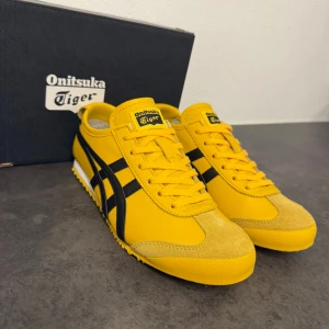 Gula Onitsuka Tiger sneakers - Säljer ett par ikoniska Onitsuka Tiger sneakers i starkt gul färg med svarta detaljer och klassiska ränder på sidorna. Skorna har snörning, platt sula med mönstrad undersida och rund tå. Materialet är en mix av skinn och mocka, vilket ger en snygg kontrast och retrovibe. Storlek 39