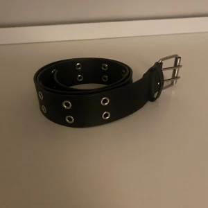 Svart skärp med silverfärgade nitar - Säljer ett svart bälte med dubbla rader silverfärgade metallöljetter och ett stilrent spänne i metall. Bältet är brett och har en cool, edgy vibe som passar perfekt till jeans eller svarta byxor. Materialet är skinnimitation och designen är enkel men ändå iögonfallande.