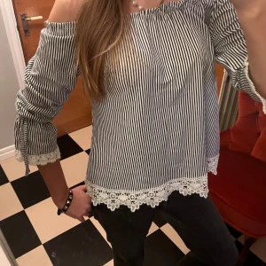 Randig offshoulder blus med spets - Säljer en mörkblå randig offshoulder blus med fina spetsdetaljer i vitt längs ärmslut och nederkant. Blusen har trekvartsärm med knyt vid ärmslut och en luftig, loose passform. Perfekt för dig som gillar romantiska detaljer och snygg stil.