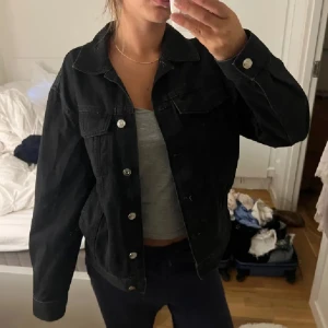 Svart jeansjacka med silverknappar - Säljer en svart jeansjacka med klassisk krage och silverfärgade knappar framtill. Jackan har två bröstfickor med lock och knapp samt långa ärmar. Perfekt för dig som gillar en tidlös och enkel stil.