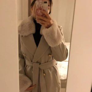 Beige kappa med fuskpäls och bälte - Säljer en beige kappa från River Island med bred krage och fuskpälsdetaljer på krage och ärmslut. Dubbelknäppt med guldknappar och tillhörande bälte i midjan. Lång modell med fickor. Avtagbar päls på kragen.  Perfekt för dig som vill ha en chic och trendig look.
