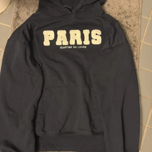 Svart hoodie med Paris-tryck - Svart hoodie med stor, fluffig text 'PARIS' i vitt på bröstet och mindre text 'QUARTIER DU LOUVRE' under. Klassisk känguruficka framtill och huva med snörning. Perfekt för dig som gillar streetstyle och vill ha en chill look.