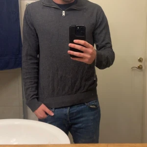 Mörkgrå stickad tröja med half zip - Mörkgrå stickad tröja med halv dragkedja vid halsen. Tröjan har en enkel och clean design med ribbade muddar och nederkant. Perfekt för lager-på-lager och funkar året runt. Snygg till både jeans och chinos.