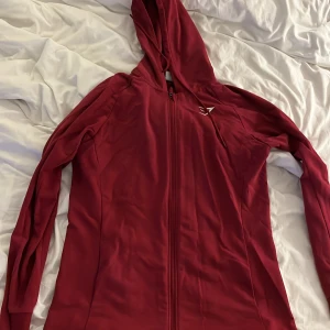 Röd hoodiejacka från Gymshark - Snygg röd hoodiejacka från Gymshark i storlek L. Jackan har hel dragkedja framtill, lång ärm och huva. Materialet är mjukt och stretchigt, perfekt för träning eller chill. Gymshark-loggan är broderad i vitt på bröstet. Passformen är normal och jackan är enkel att matcha.