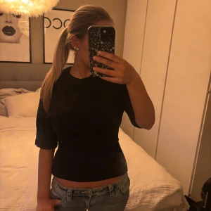 Svart omlottopp med knyt i ryggen - Säljer en svart omlottopp med snygg knytning i ryggen. Toppen har korta ärmar och en tight passform som framhäver midjan. Materialet känns mjukt och stretchigt, perfekt att styla med jeans eller kjol. Enkel och stilren design som ger en cool vibe.