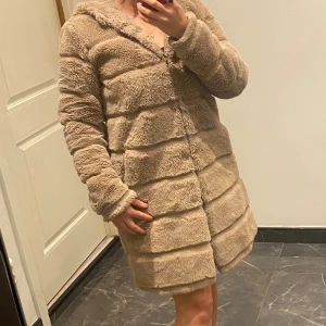 Beige pälsjacka med huva - Säljer en supermysig beige pälsjacka med huva. Jackan har ett fluffigt och mjukt material, långa ärmar och en rak passform som går ner till låret. Snygga horisontella sömmar ger en cool struktur. Perfekt för kalla dagar när du vill vara både varm och stylish.