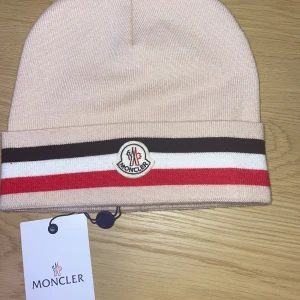 Beige Moncler mössa med ränder - Snygg beige mössa från Moncler med klassiska broderade loggan framtill. Mössan har breda randiga detaljer i brunt, vitt och rött längs kanten. Tillverkad i mjuk stickad ull som känns lyxig och varm. Perfekt för dig som vill ha en stilren accessoar med premiumkänsla.