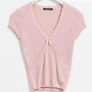 Ljusrosa stickad bolerotopp med rosett - Supersöt ljusrosa stickad bolerotopp med korta ärmar och v-ringning. Toppen har en liten rosett framtill och vågiga kanter längs halsringningen. Perfekt för att addera en gullig touch till din outfit. Liten i storleken, passar mig som har storlek S.