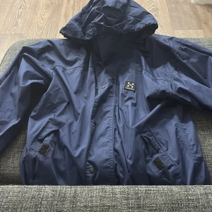 Blå vindjacka från Haglöfs XS - Säljer en blå vindjacka från Haglöfs i storlek XS. Jackan har huva, dragkedja och tryckknappar framtill samt två fickor med kardborre. Materialet är lätt och syntetiskt, perfekt för blåsiga dagar. Klassisk Haglöfs-logga på bröstet. Passar xs-m 