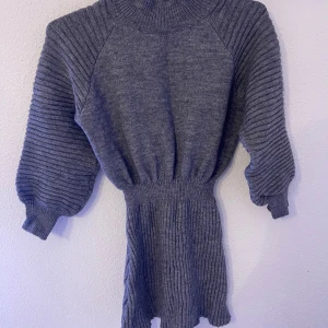 Grå ribbad stickad klänning - Mysig grå stickad klänning med ribbade detaljer och ballongärmar. Hög hals och markerad midja ger en snygg siluett. Perfekt för dig som gillar en enkel men trendig look under kyligare dagar.