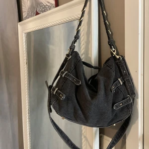 Svart denim axelväska med spännen - Grå jeans axelväska med flera dekorativa spännen och metallringar. Väskan har en mjuk form och justerbar axelrem. Väskan är från Zara och nypris är 700kr 💗🫶🏼