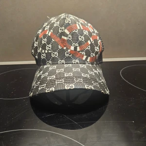 Keps med orm och GG-mönster - Snygg svart keps med klassiskt GG-mönster över hela kepsen och en röd-vit orm på framsidan. Justerbar rem baktill i grönt och rött. Kepsen har mesh på sidorna och baksidan för extra ventilation. Perfekt för dig som vill sticka ut med en lyxig accessoar.