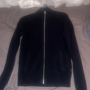 Svart stickad kofta från Jack & Jones - Mörk, ribbstickad kofta från Jack & Jones i svart med hel dragkedja framtill och två fickor. Perfekt för lager-på-lager och har en clean, enkel look. Skön och mjuk känsla tack vare det stickade materialet.