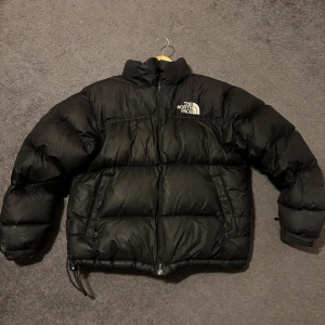 Svart pufferjacka från The North Face - Säljer en svart pufferjacka från The North Face i storlek M. Jackan har en hög krage, två sidofickor med dragkedja och tydlig logga på bröstet och ryggen. Den är vadderad och har en loose passform, perfekt för kalla dagar. Allmänt så är den i bra skick. 