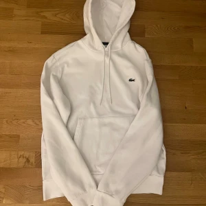 Lacoste hoodie - Vit hoodie från Lacoste med klassisk krokodillogo på bröstet. Tröjan har huva med snörning och en stor magficka framtill. Mjuk bomullskvalitet och långärmad modell som är perfekt för chill dagar. Enkel och stilren design som funkar till allt.