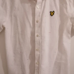Vit kortärmad skjorta från Lyle & Scott Junior - Vit kortärmad skjorta från Lyle & Scott Junior med klassisk krage och knappar framtill. Skjortan har små färgstänk i blått, rött och gult samt den ikoniska broderade gula örnen på bröstet. Tillverkad i mjuk bomull, perfekt för en clean och fräsch look.