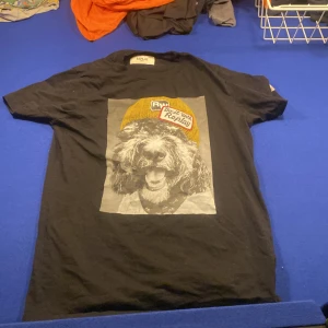 Svart t-shirt Replay med hundtryck - Svart t-shirt från Replay i storlek M med ett stort tryck på framsidan av en hund med gul mössa och texten 'Do it with Replay'. Klassisk rund hals och korta ärmar. Perfekt för dig som gillar coola och unika motiv.