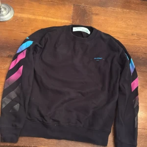 Off-White svart sweatshirt med färgglada detaljer - Svart sweatshirt från Off-White med ikoniska pilar och färgblock i blått, rosa och lila på ärmar och rygg. Crewneck-modell med tryckta detaljer och klassisk passform. Perfekt för dig som gillar streetwear och vill sticka ut.