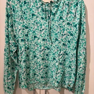 Blommig blus från Pernilla Wahlgren XS - Supersöt blommig blus från Pernilla Wahlgren i vitt och grönt. Långärmad modell med knyt i halsen och små volangdetaljer. Tunt och mjukt material, perfekt för vår och sommar. Guldig knappdetalj vid ärmslutet ger extra touch.