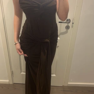 Brun långklänning från Boohoo - Supersnygg brun långklänning från Boohoo med omlott-detalj och drapering i midjan. Klänningen är ärmlös och har en tight passform som framhäver figuren. Materialet är mjukt och stretchigt, perfekt för dig som vill ha en chic och trendig look.