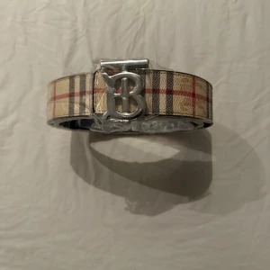Rutig Burberry bälte med silverdetalj - Snyggt bälte från Burberry med klassiskt rutigt mönster i beige, rött och svart. Bältet har ett stort silvrigt spänne med Burberrys ikoniska B-logga. Perfekt accessoar för att lyfta din outfit.