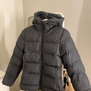 Svart pufferjacka med huva - Svart pufferjacka med huva och dragkedja framtill. Jackan har två sidofickor med dragkedja och justerbar snörning vid huvan. Den quiltade designen ger en schysst volym och värme, perfekt för kalla dagar. Enkel och stilren look som funkar till det mesta. Se de två sista bilderna för skadorna men de första är lagade och synd inte medan märket har blivit lite skrynkligt men är ej något man tänker på. Kostade över 2000 kr ny, skriv vid frågor 