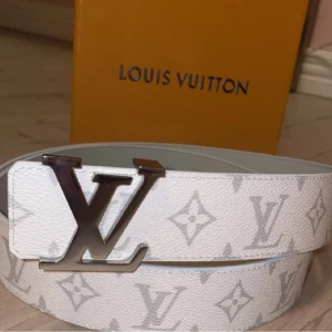 Vit Louis Vuitton monogram bälte - Snyggt vitt bälte från Louis Vuitton med klassiskt monogrammönster i ljusgrått och stor LV-spänne i silver. Bältet är gjort i präglat skinn och har en exklusiv känsla som lyfter vilken outfit som helst.