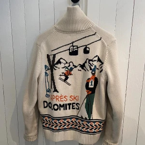 Stickad kofta Morris Après Ski Dolomites - Sällsynt & unik retroinspirerad stickad kofta från Morris med hög krage och dragkedja. Beige bas med färgglada skidmotiv, texten 'Après Ski Dolomites' och mönstrad kant. Quiltad insida för extra värme. Perfekt för dig som gillar vintagevibbar och vintersport.
