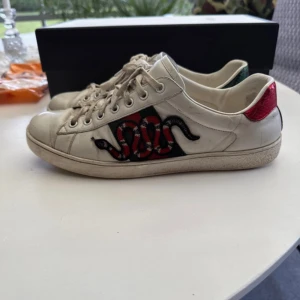 Gucci sneakers med orm broderi - Snygga vita Gucci sneakers i skinn med broderad röd och svart orm på sidan. Skorna har snörning, platt sula och detaljer i rött och grönt på hälen. Ikonisk design som sticker ut och passar perfekt till streetwear. Kommer med originalförpackning och dustbag.