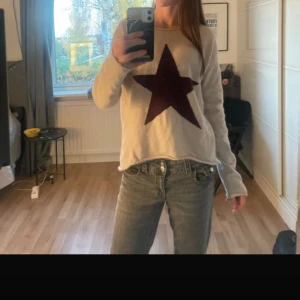 Beige stickad tröja med stjärna Hunkydory - Supermjuk beige stickad tröja från Hunkydory med stor mörklila stjärna framtill. Tröjan har rund halsringning och långa ärmar, perfekt för lager på lager. Snygg till jeans eller kjol för en chill vibe.