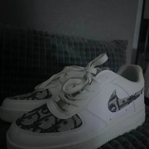 Nike Air Force 1 med svartvitt mönster - Säljer ett par vita Nike Air Force 1 sneakers med unikt svartvitt mönster på swoosh-loggan och tåpartiet. Skorna har klassisk rund tå, snörning och platt sula. Tillverkade i skinn och syntet, perfekta för dig som vill sticka ut med en clean men ändå personlig stil.