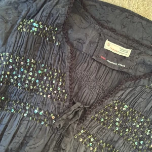 Mörkblå kortklänning med paljetter från Odd Molly - Supersöt mörkblå kortklänning från Odd Molly med glittriga paljetter på bröstet och midjan. Klänningen har knytning framtill, smockade detaljer och trekvartsärm. Materialet är mjukt och har en lätt struktur som ger en bohemisk vibe.